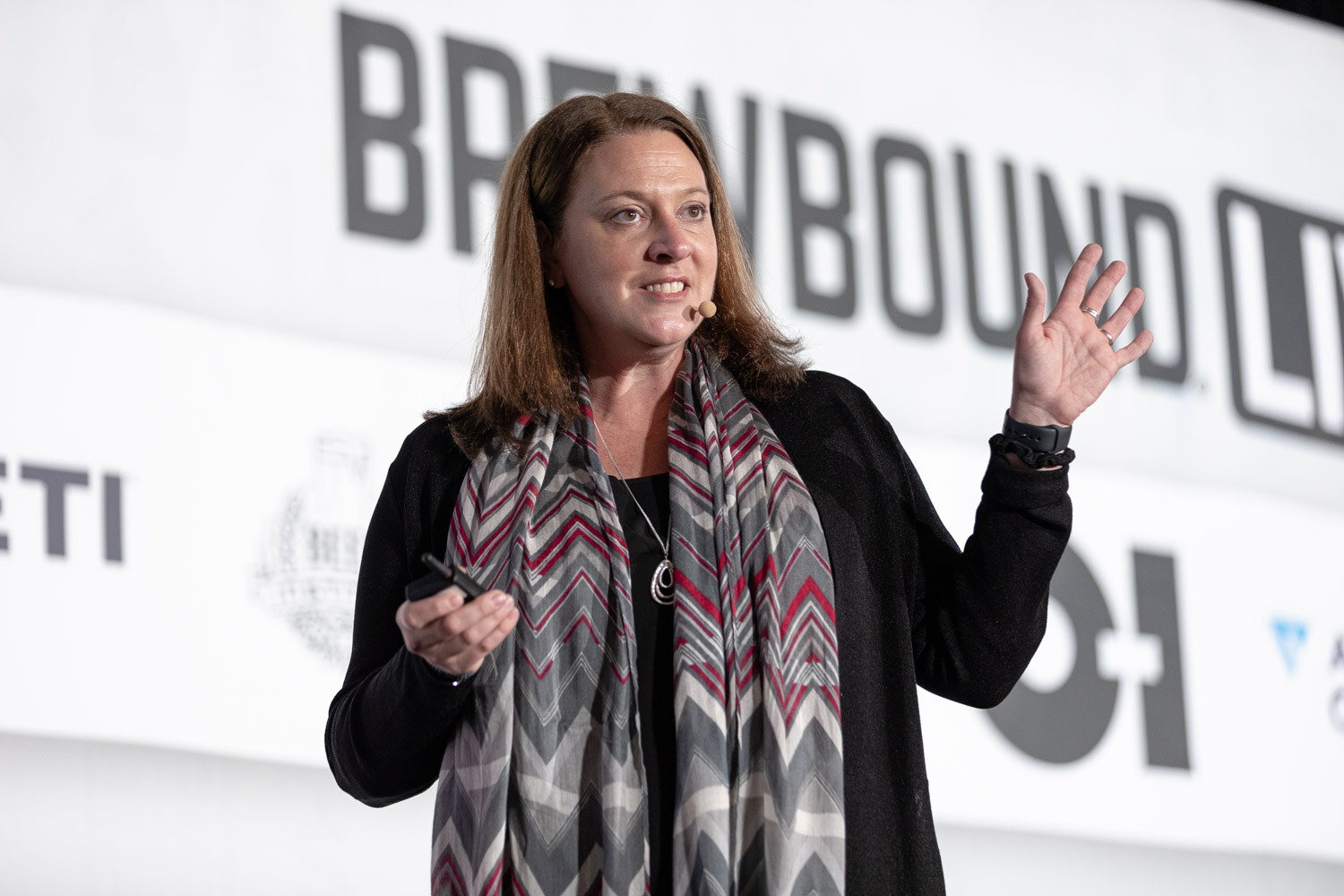 A woman in a black cardigan and gray patterned scarf holds a clicker and raises one hand in front of a Brewbound Live banner.