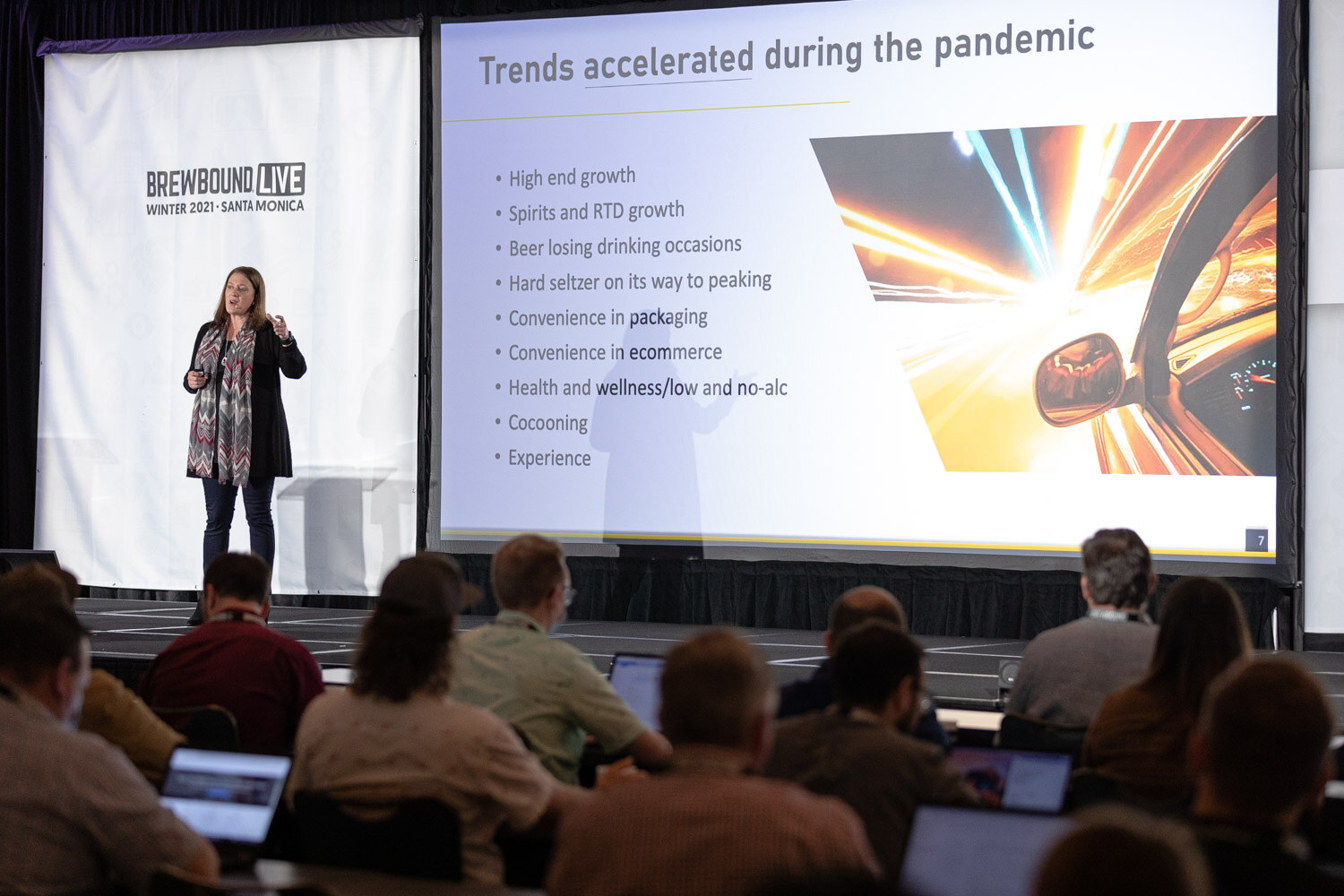 A woman stands on stage beside a screen listing trends accelerated during the pandemic, with attendees and laptops in the foreground.