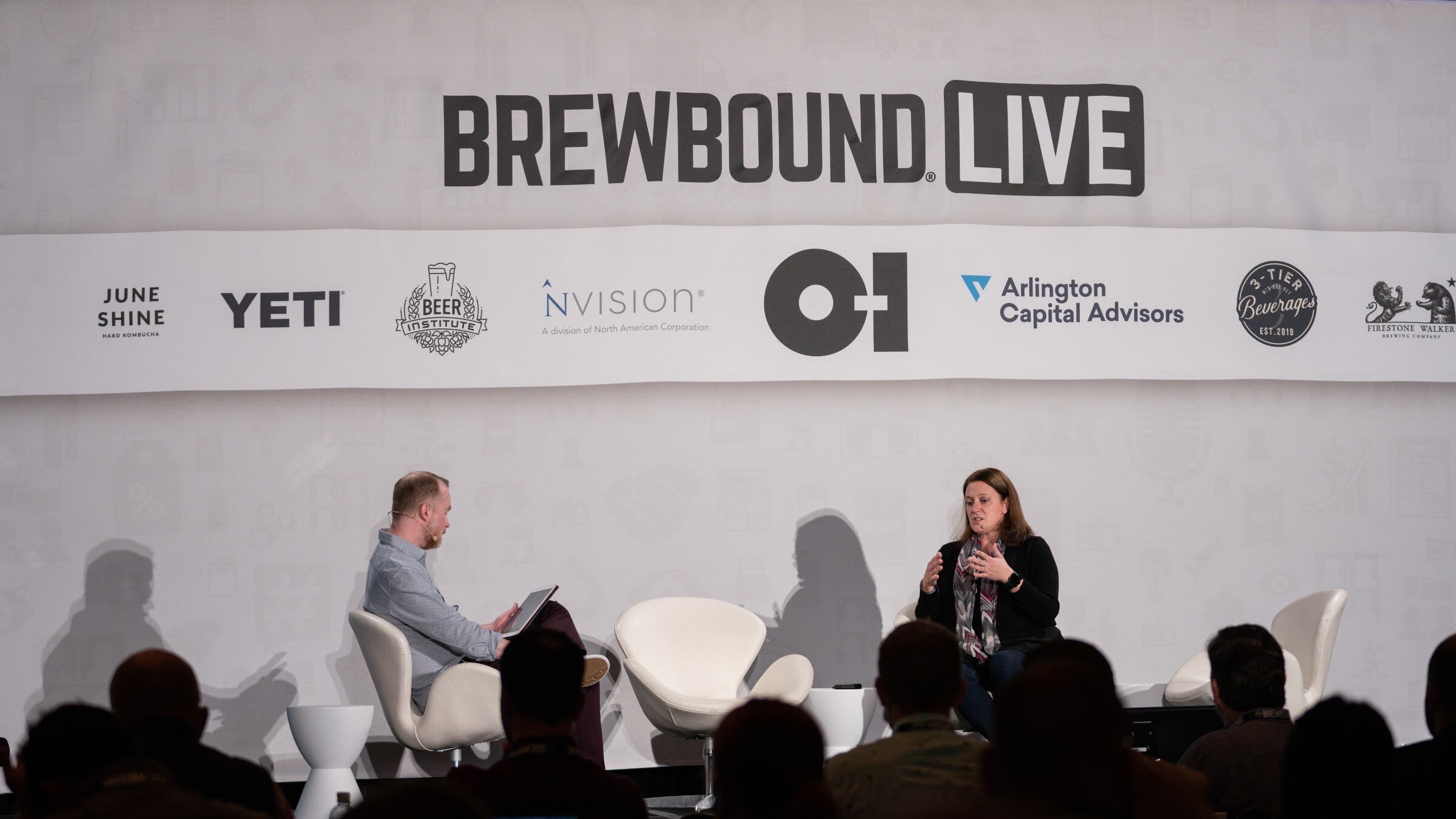Two people sit on a wide stage under a Brewbound Live banner with sponsor logos while the audience watches in silhouette.