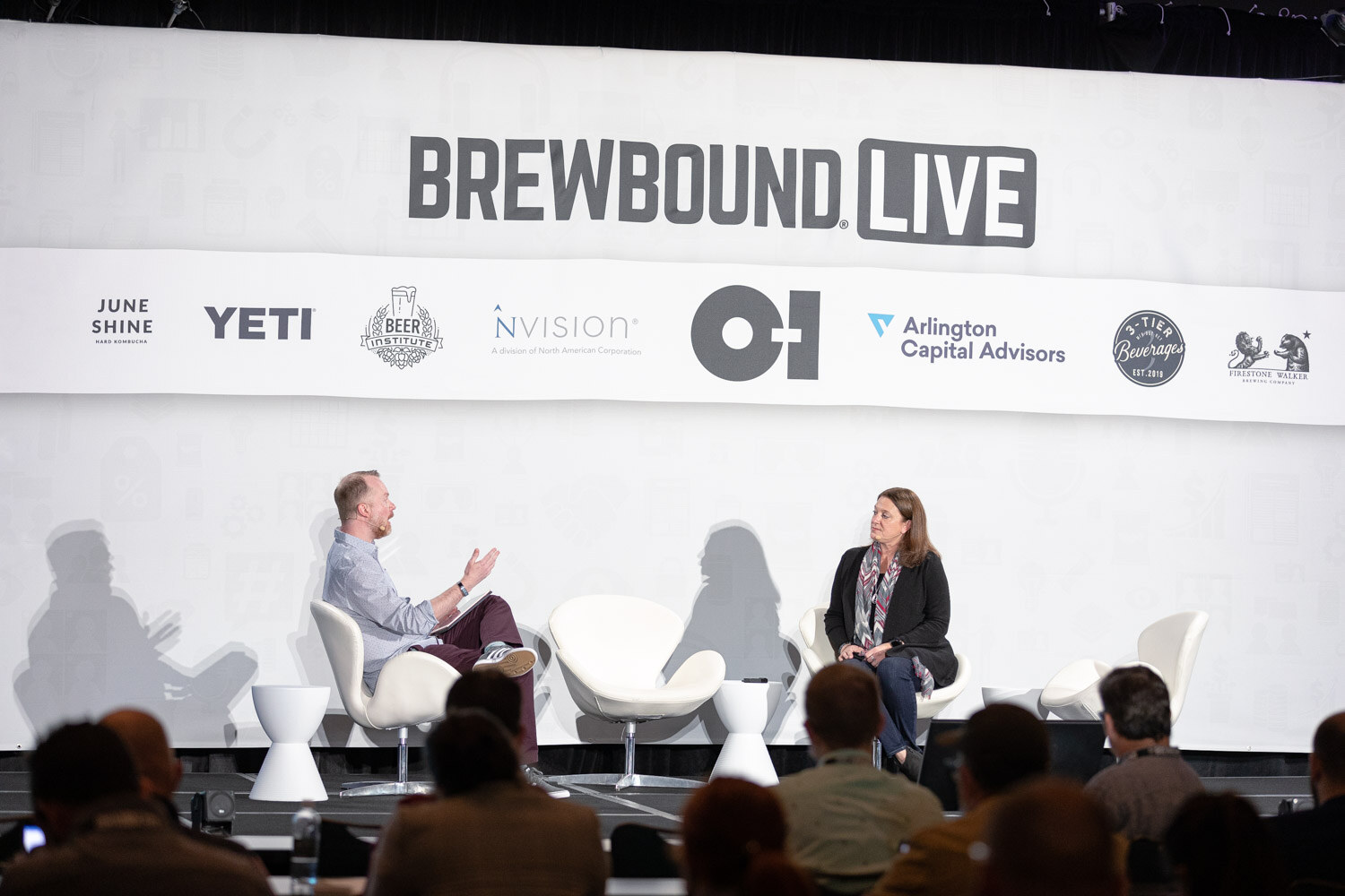 Two people sit on a wide stage with a Brewbound Live sponsor banner behind them, with audience members in the foreground.
