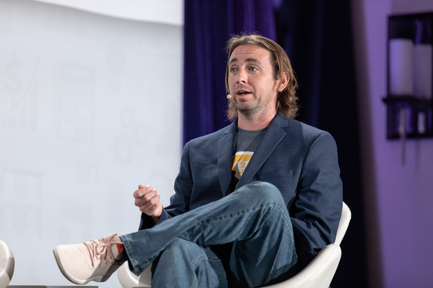 A man with shoulder-length hair in a navy blazer sits cross-legged in a white chair and speaks.