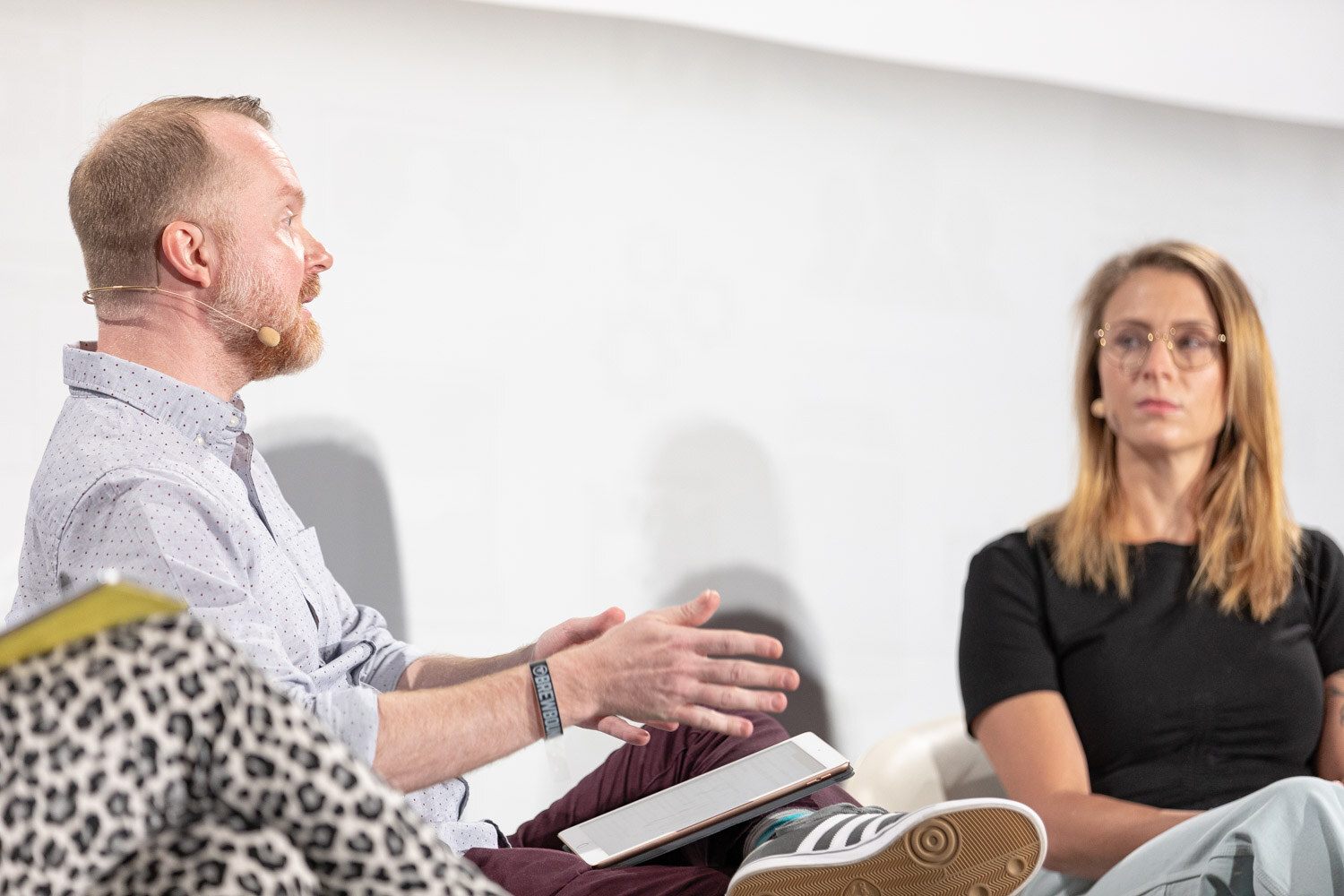 A bearded man in a light shirt gestures with an iPad on his lap as a woman in a black top listens.