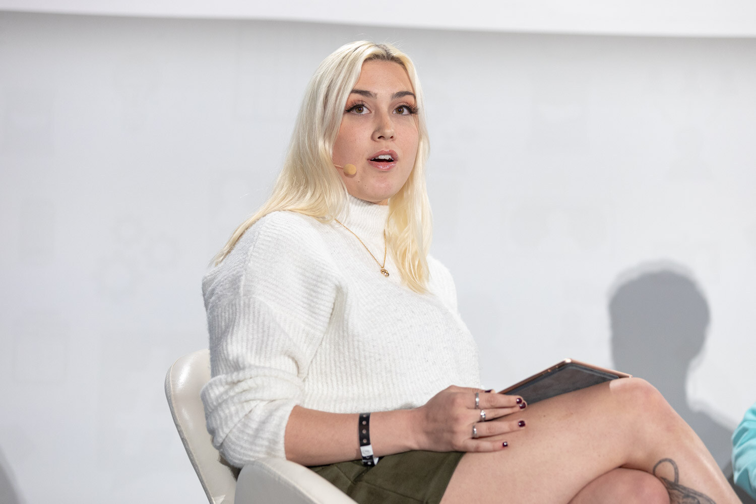 A woman with long blonde hair in a white sweater holds a tablet on her lap and speaks while seated.