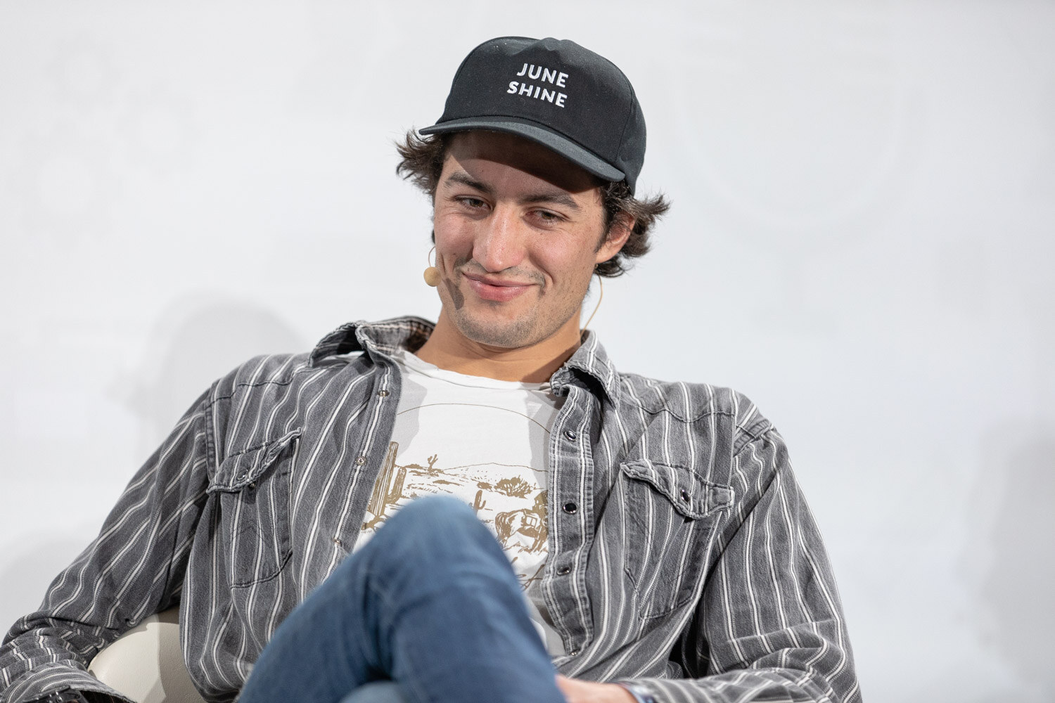 A man in a black JuneShine cap and striped shirt sits with his legs crossed, smiling.