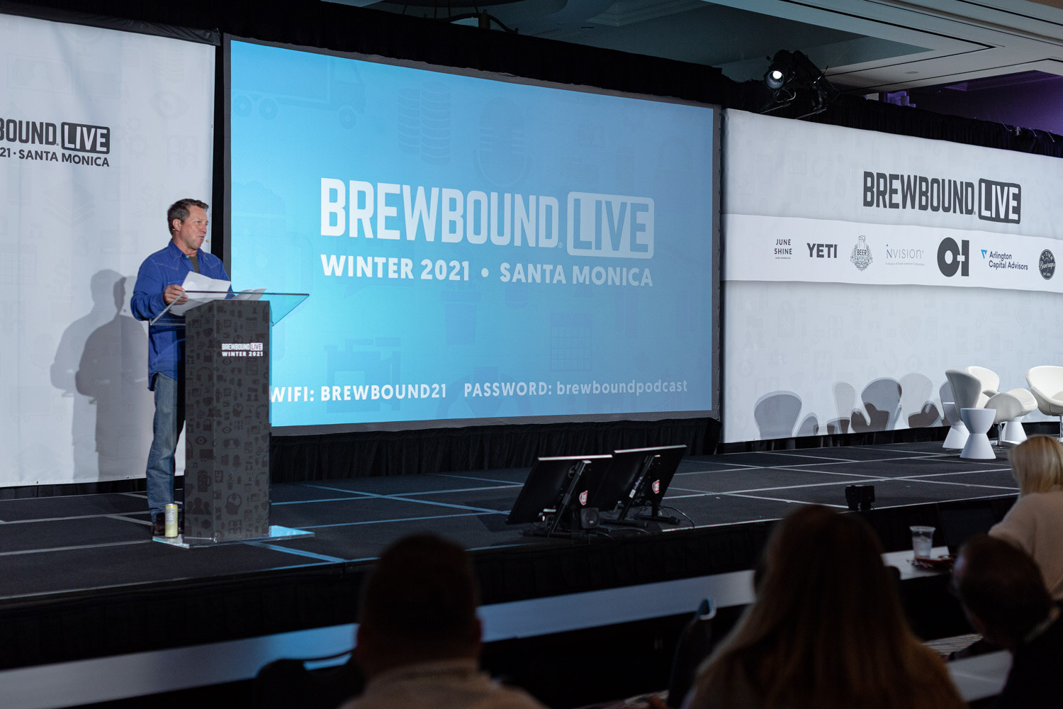 A man in a blue denim shirt at a podium beside a large blue Brewbound Live Winter 2021 Santa Monica screen showing the wifi details.