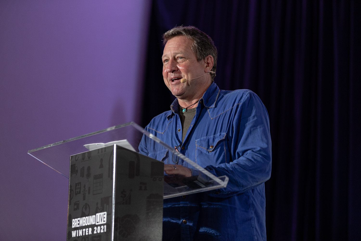 A man in a blue denim shirt smiles as he speaks at a clear podium on a purple-lit stage.