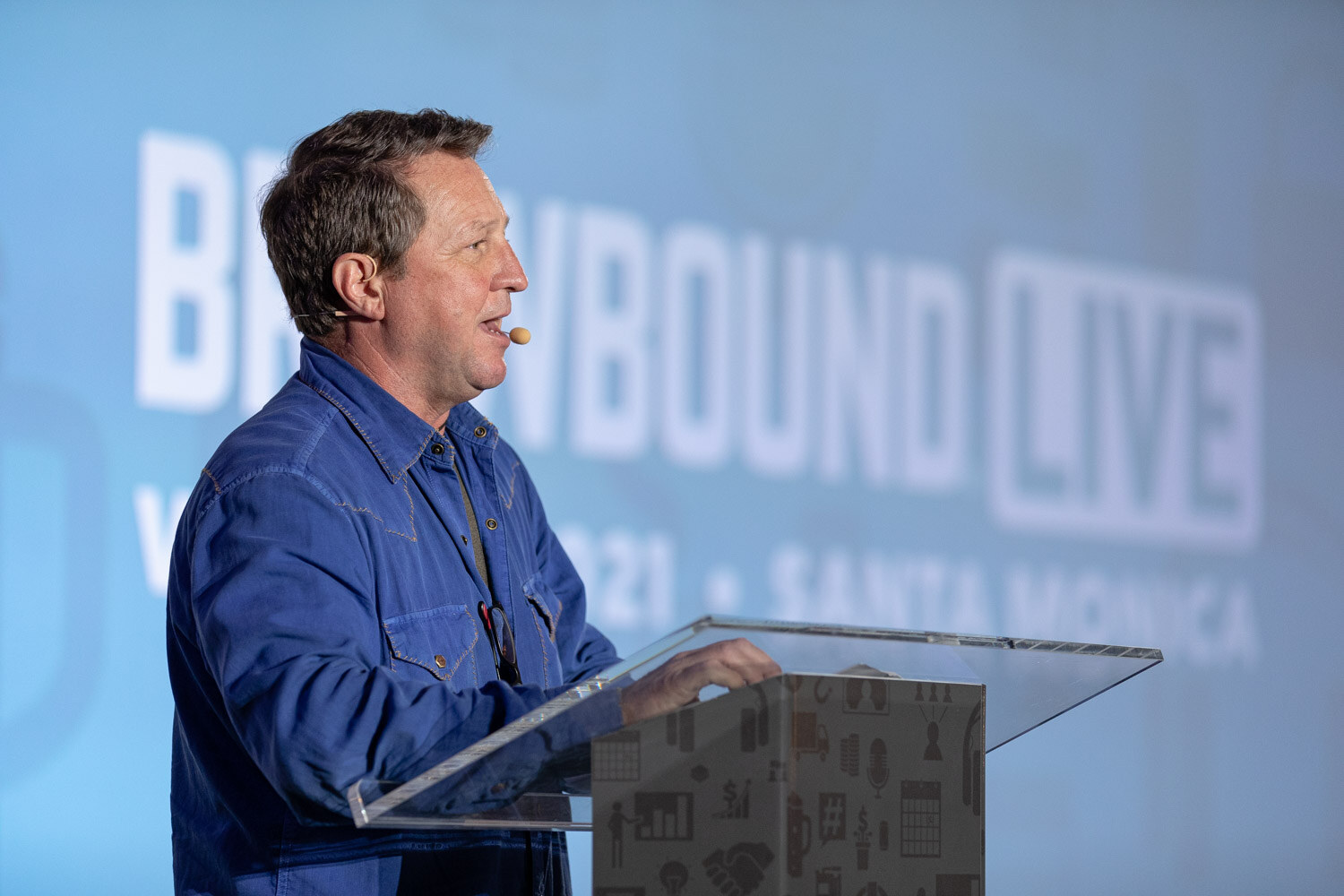 Side view of a man in a blue shirt speaking at a podium in front of a blurred blue Brewbound Live screen.
