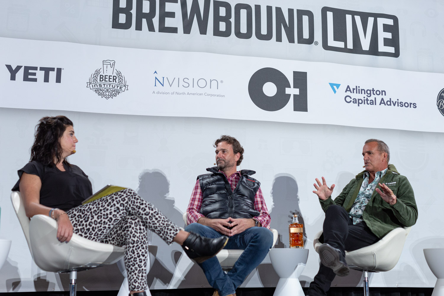 A woman in leopard print pants sits beside two men, one in a black vest and one in a green jacket, in front of a Brewbound Live banner.