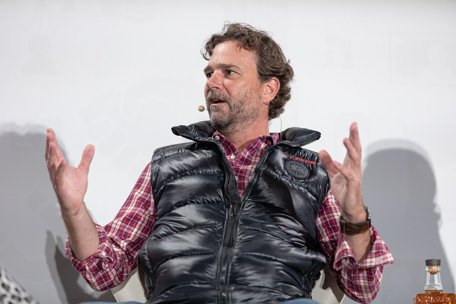 A man in a black puffer vest over a plaid shirt raises both hands while speaking, with a bottle of whiskey at the edge of the frame.