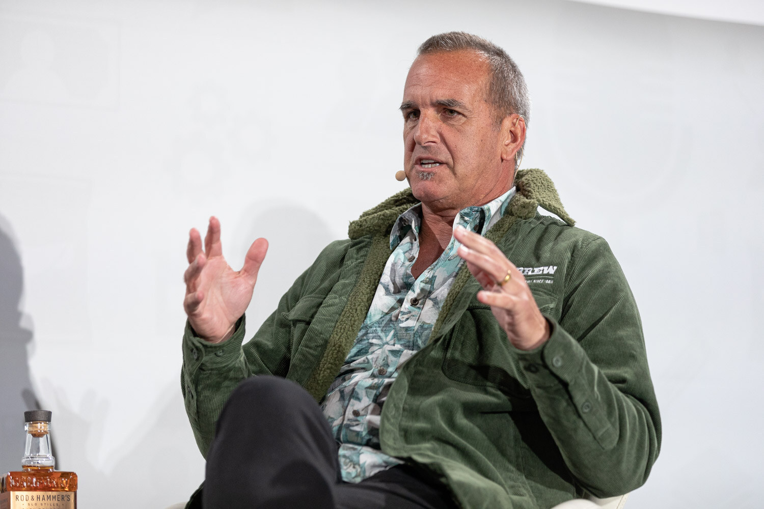 A man in a green fleece jacket over a patterned shirt gestures with both hands next to a whiskey bottle.