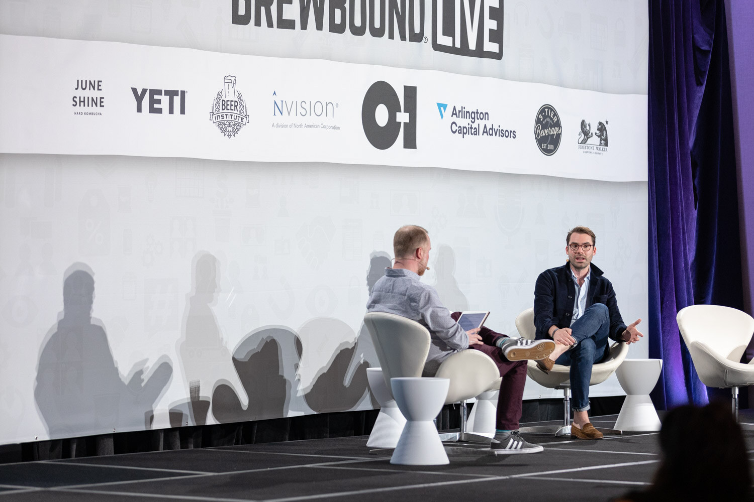 Two men sit in white chairs on a stage in front of a banner with JuneShine, YETI, Beer Institute and O-I logos.