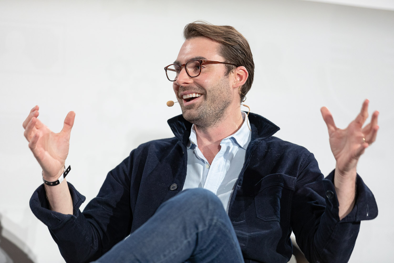 A man in glasses and a navy jacket smiles broadly with both hands raised.