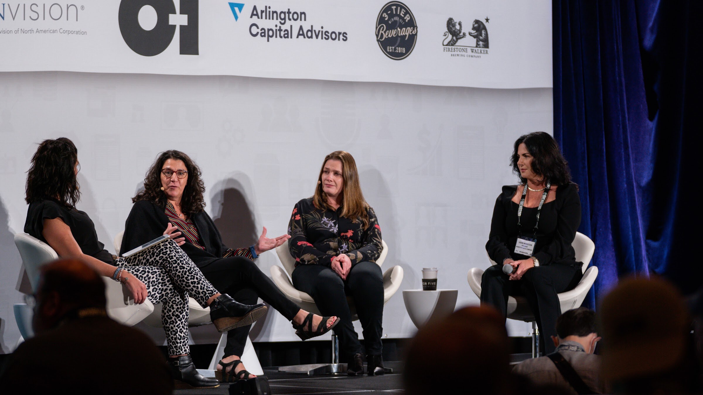 Four women sit in white chairs on a stage under a banner with sponsor logos including O-I and Arlington Capital Advisors.