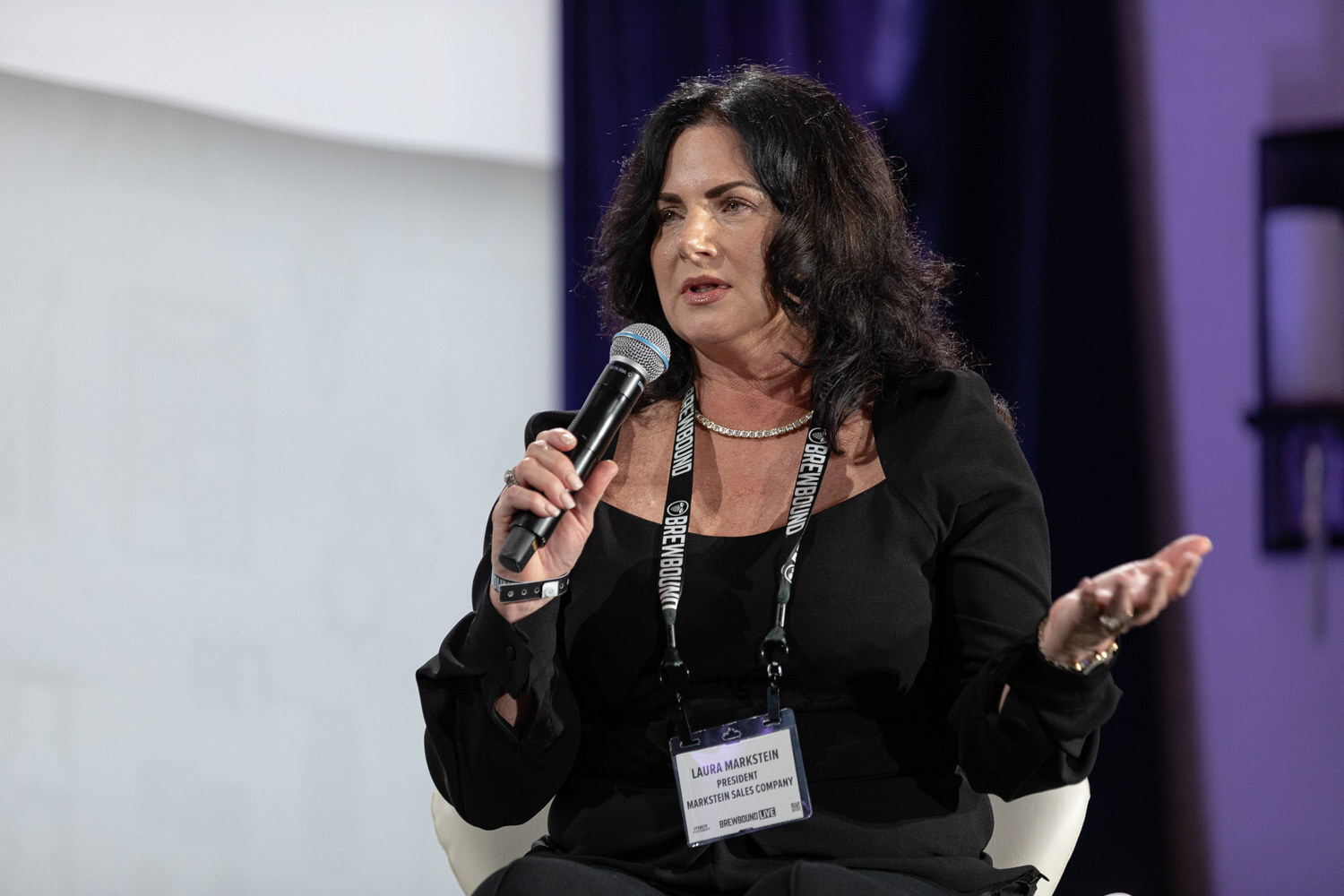 A woman with dark hair and a lanyard holds a microphone and gestures with the other hand.