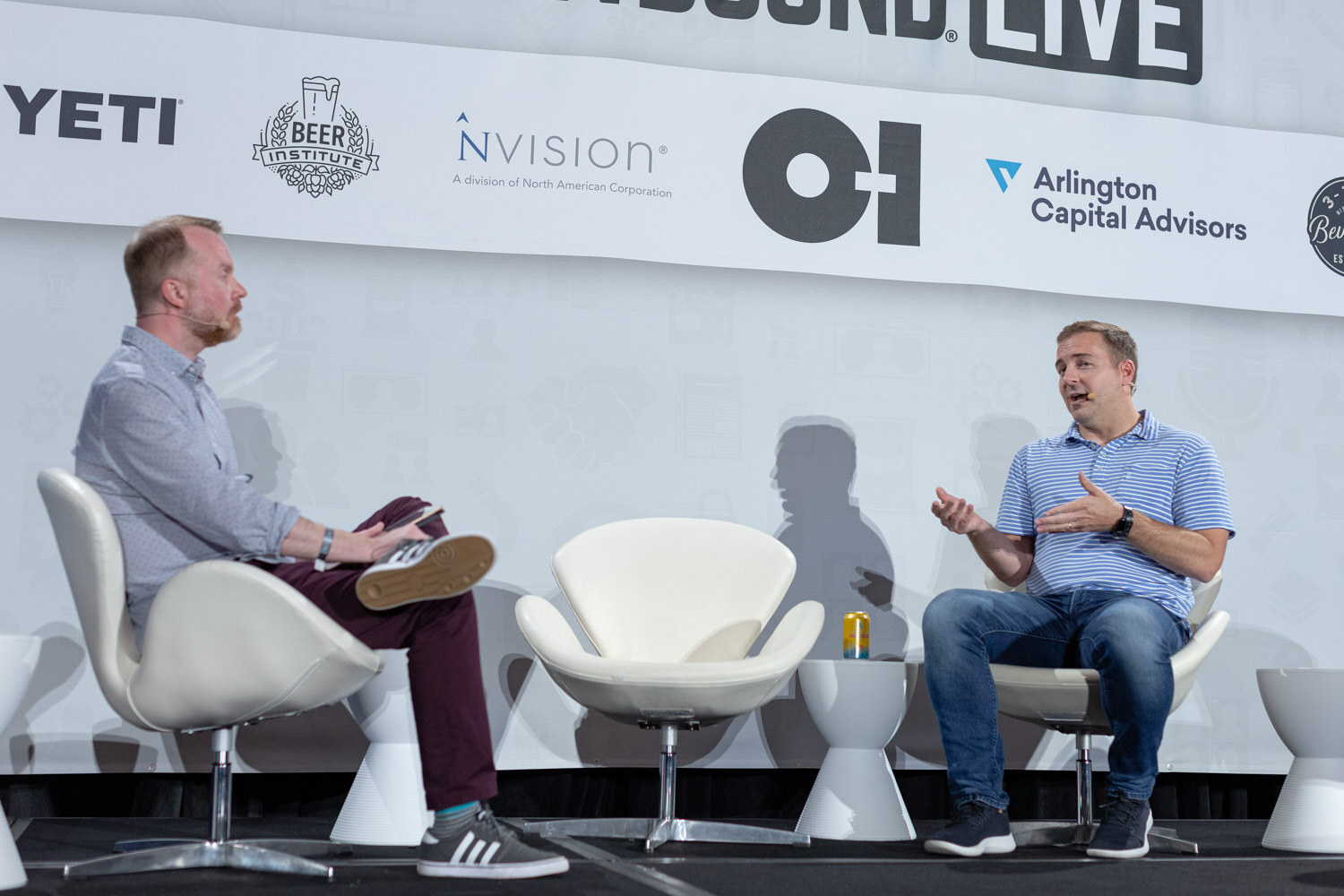 Two men sit on a wide stage, one in a striped blue polo gesturing, with a can on a stool between them under a Brewbound Live banner.