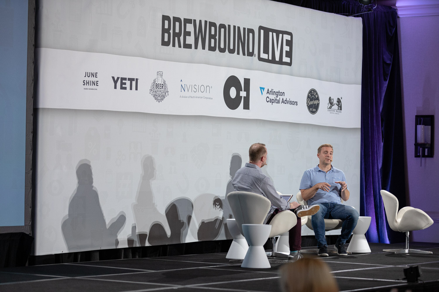 Two men sit in white chairs on a stage in front of a Brewbound Live banner with sponsor logos.