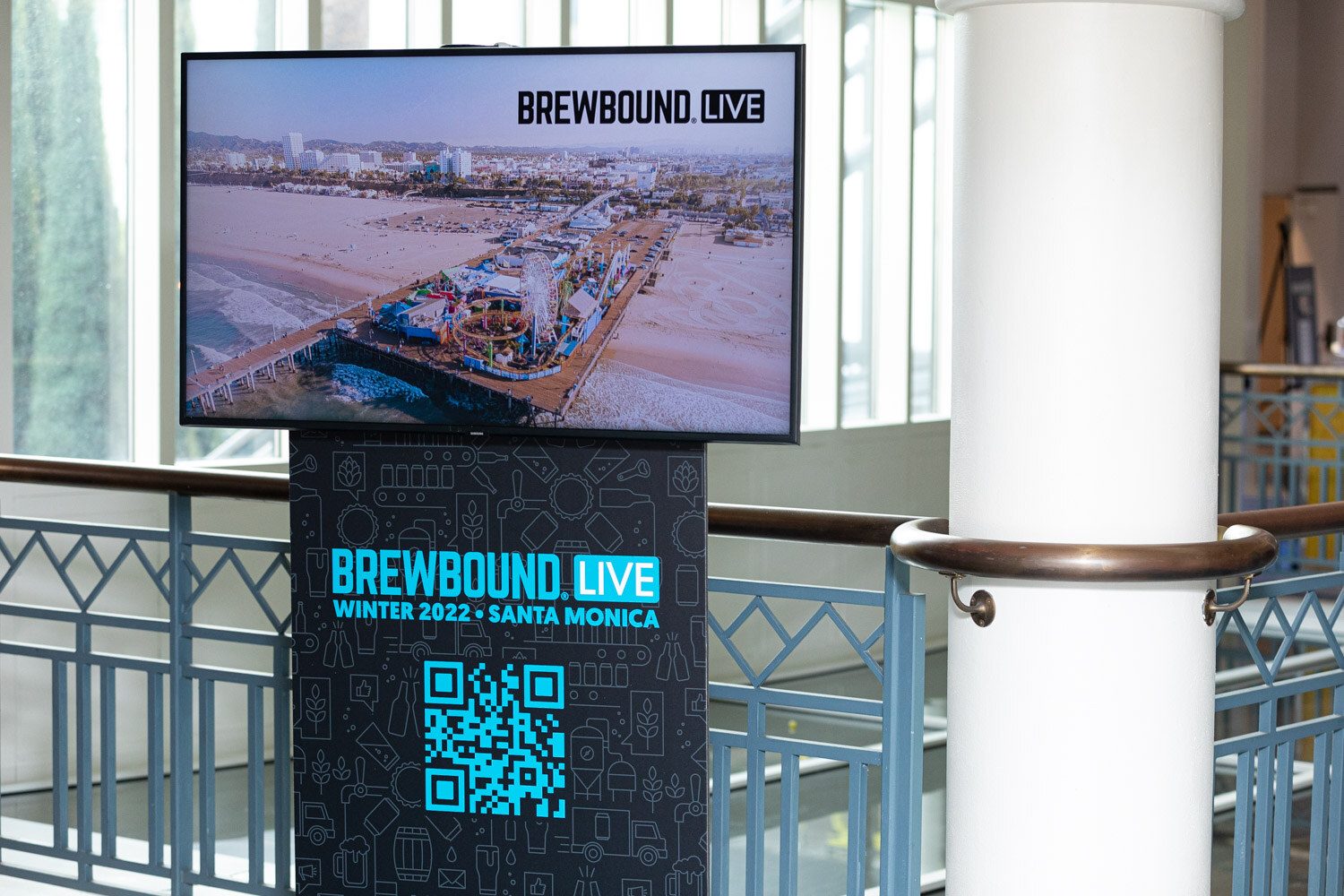 A monitor showing the Santa Monica pier above a black stand reading Brewbound Live Winter 2022 Santa Monica with a QR code.