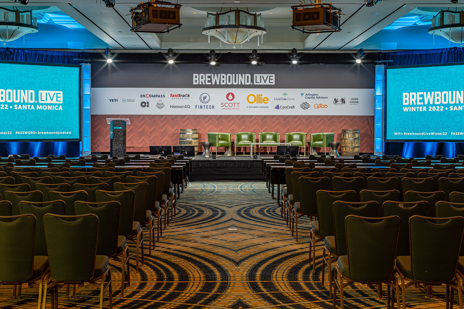 Wide view of a ballroom with rows of chairs facing a stage with green armchairs and a Brewbound Live backdrop with sponsor logos.