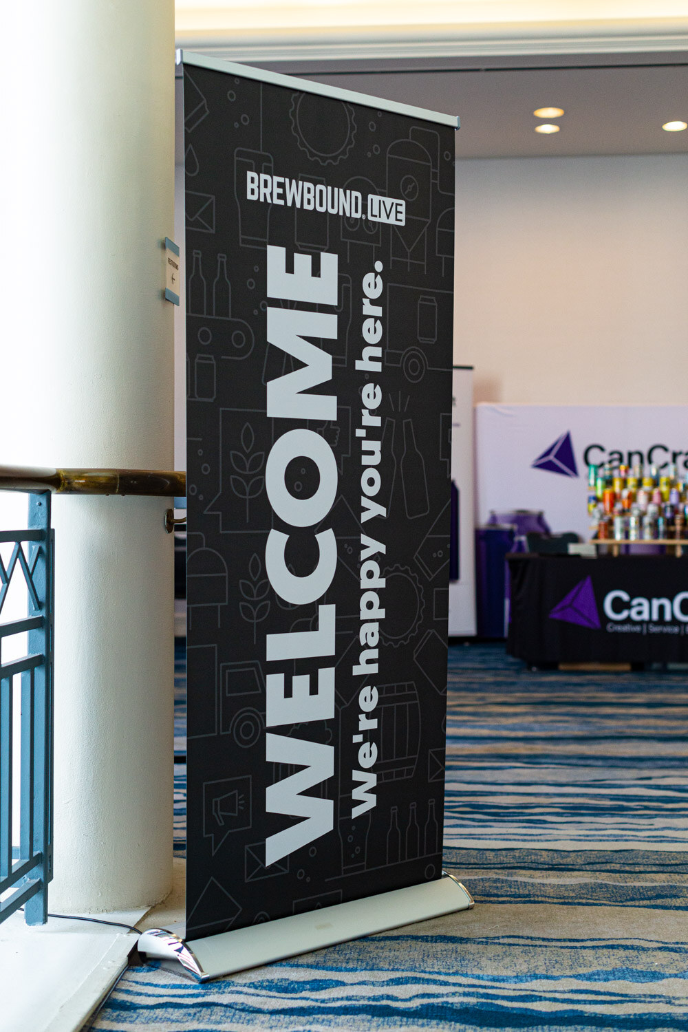 A tall black banner reading Welcome, we're happy you're here beside a column, with a CanCraft booth in the background.