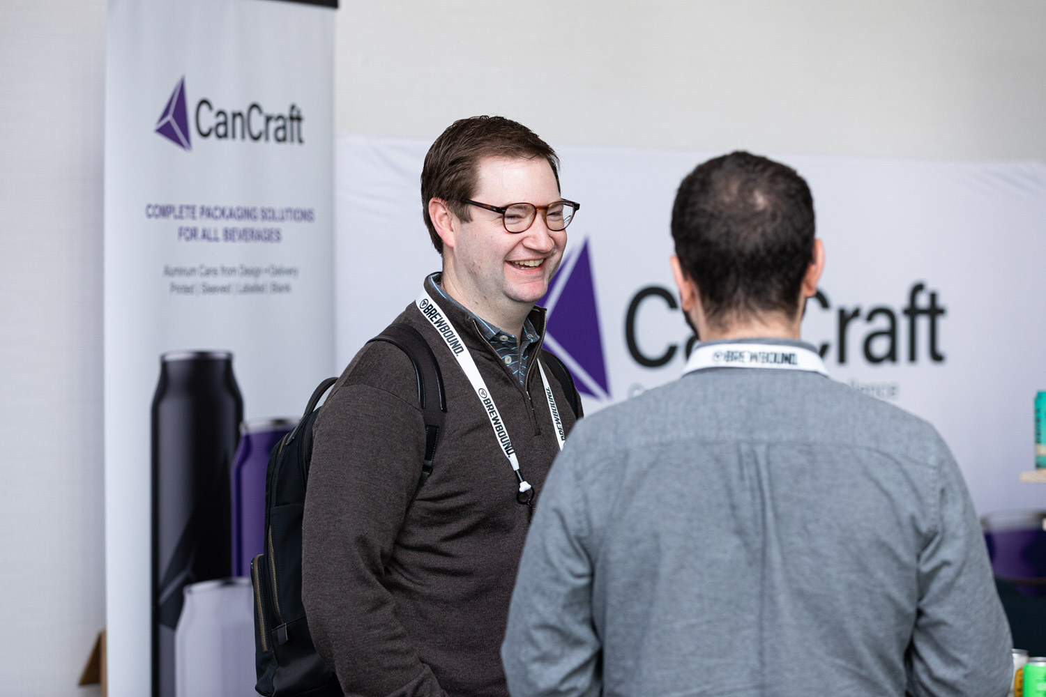A smiling man with glasses and a lanyard stands in front of a white CanCraft banner talking with another attendee.