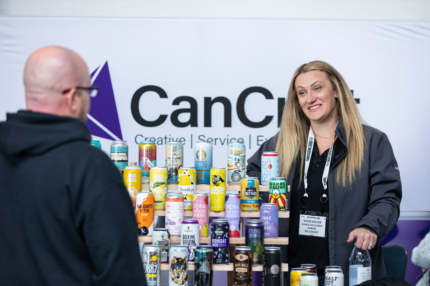 A woman with long blonde hair and a lanyard stands behind a display of colorful cans in front of a CanCraft banner.