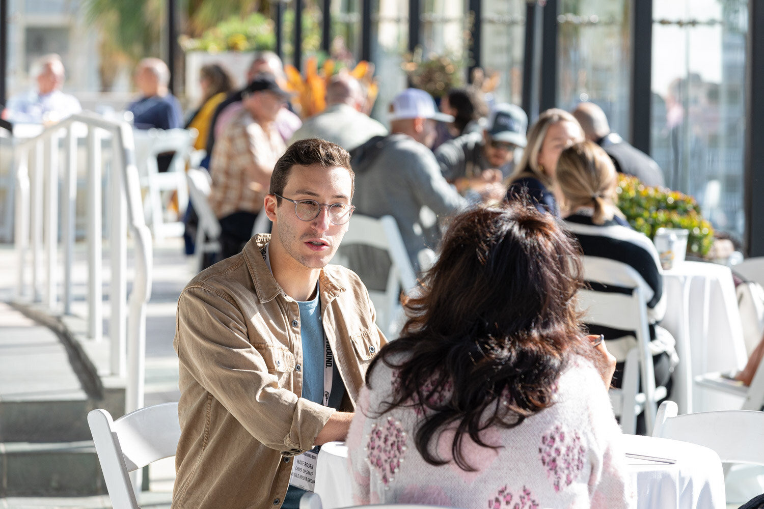 A man in a tan jacket and a woman in a pink patterned sweater sit at a white terrace table with other diners behind them.