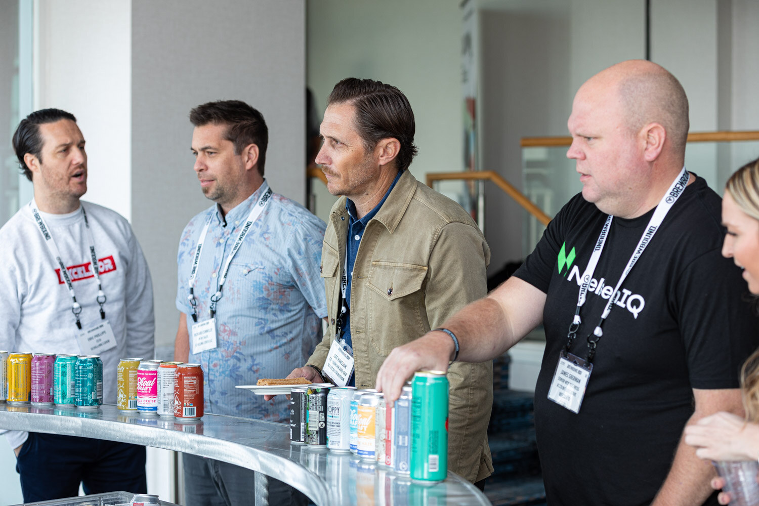 Four men with lanyards stand at a curved counter lined with colorful cans, one in a NielsenIQ T-shirt.