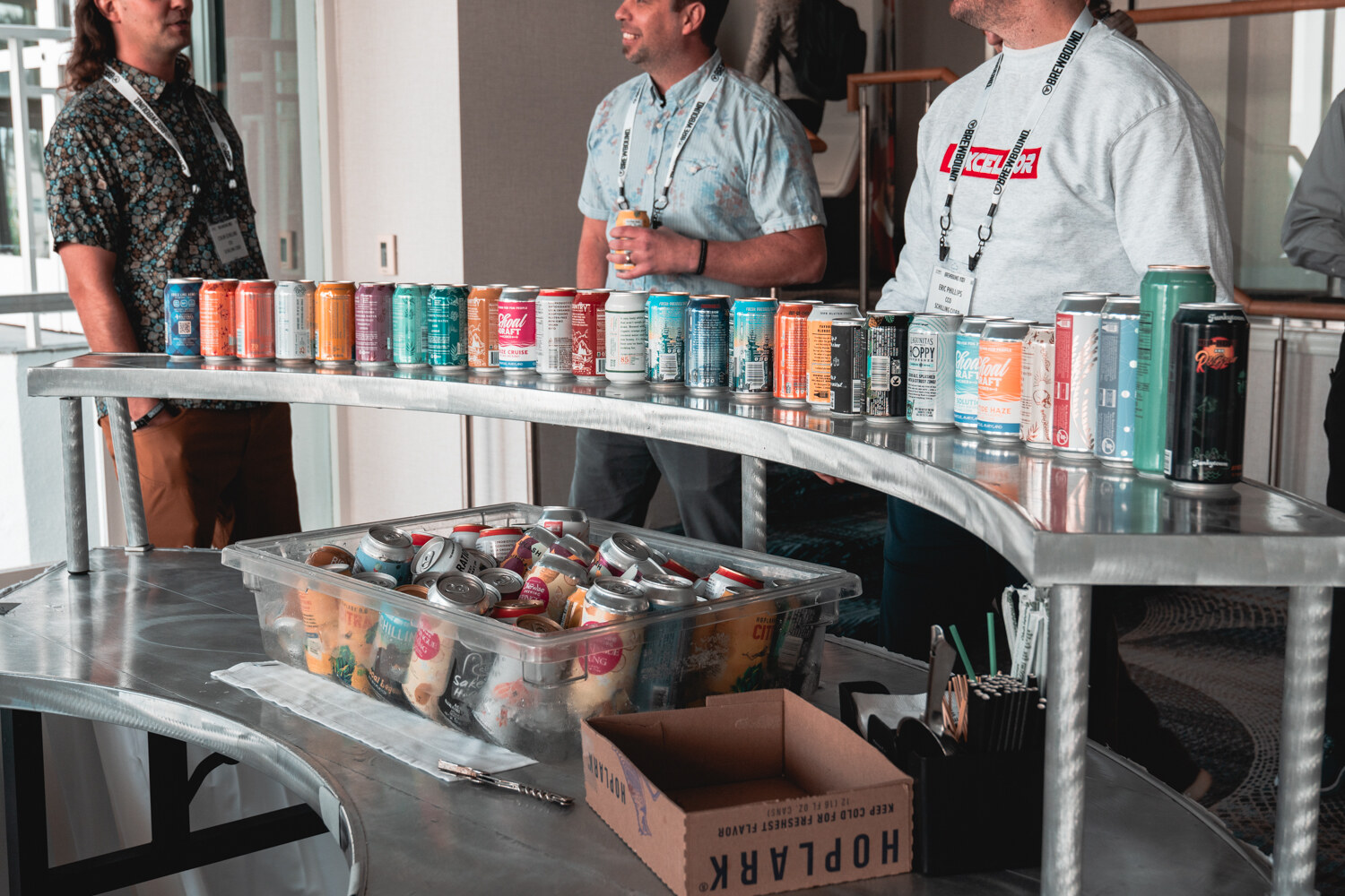 Two men with lanyards stand behind a curved silver counter displaying dozens of colorful beverage cans, with a Hoplark box below.