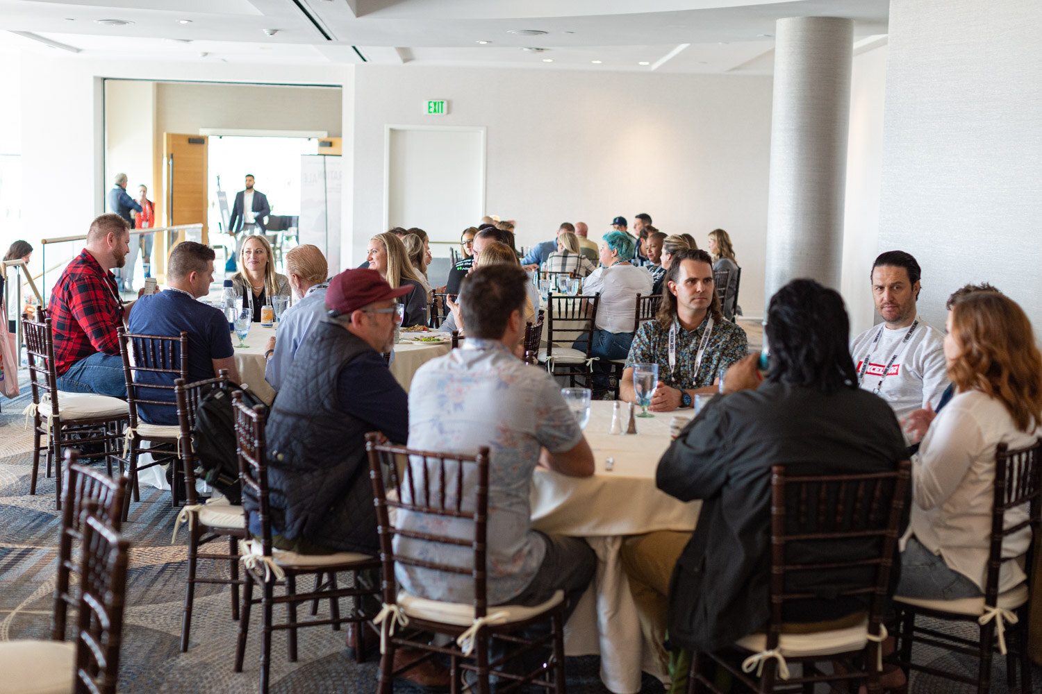 Wide view of attendees seated at round tables with gold cloths and dark chairs in a bright banquet room.