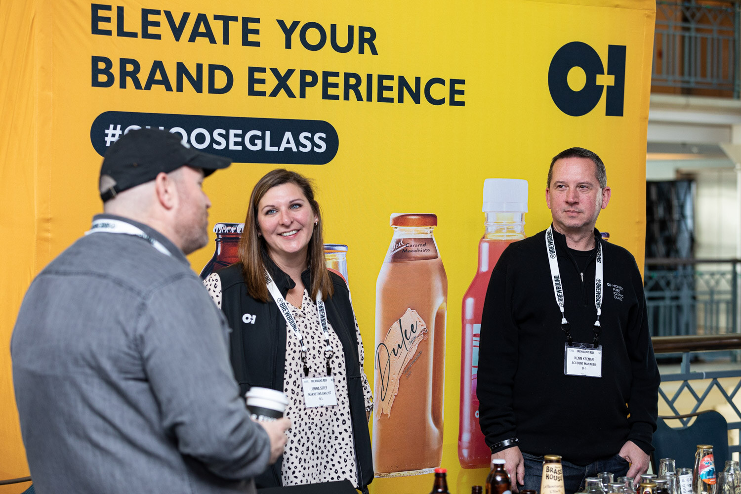 A woman in a patterned top and a man in black stand at a yellow banner reading Elevate Your Brand Experience #ChooseGlass with glass bottles in front.