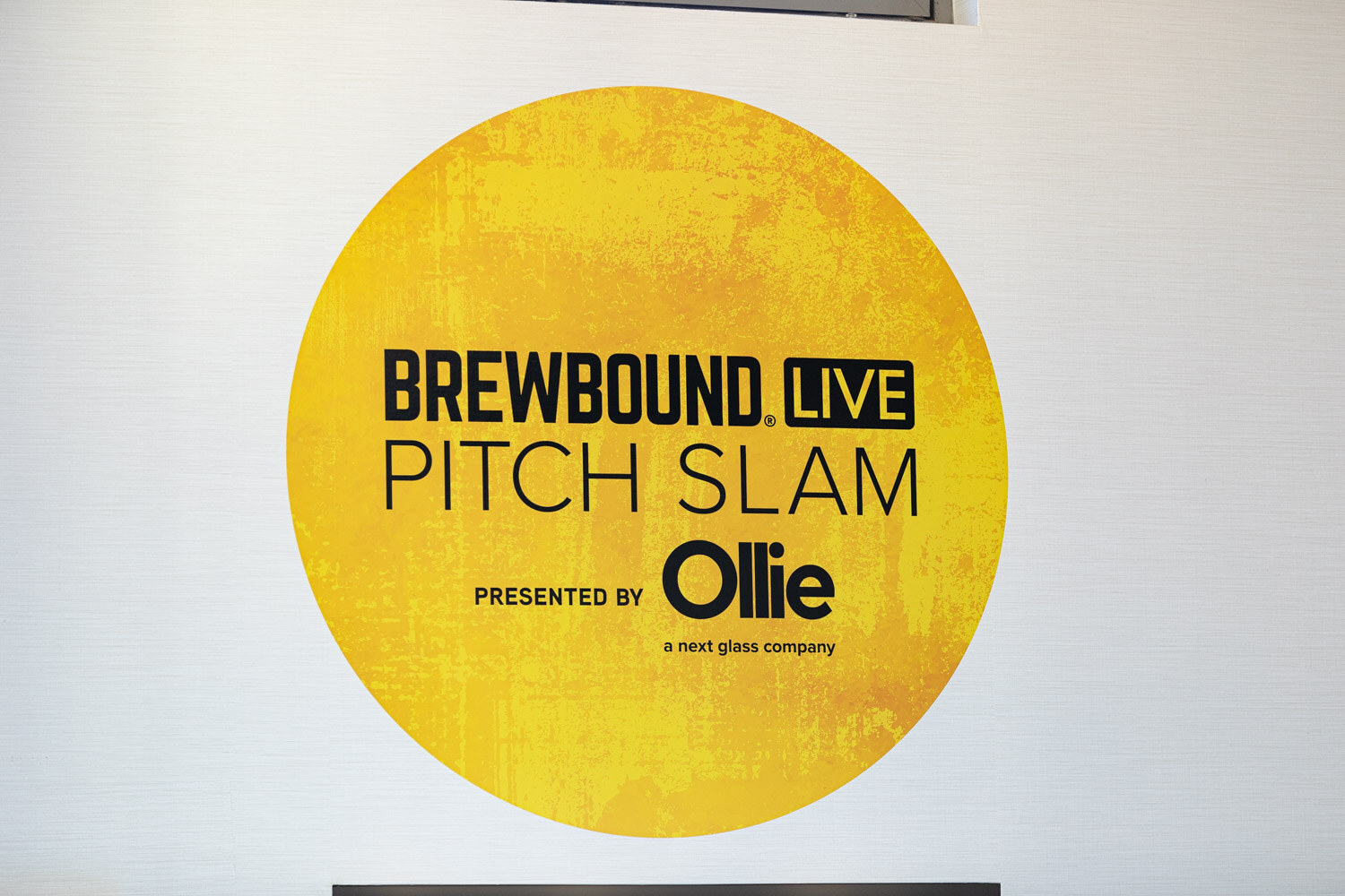 A yellow disc on a white wall reading Brewbound Live Pitch Slam presented by Ollie.