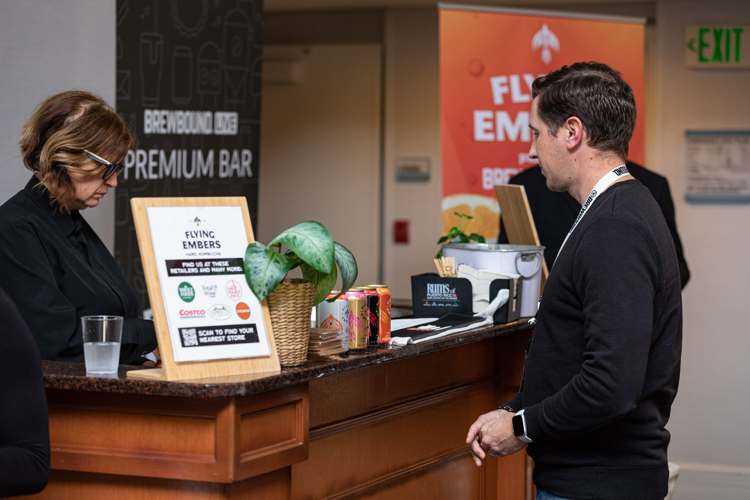 A man in a black sweater pours from a can at a wooden counter marked Premium Bar with a Flying Embers sign and a potted plant.
