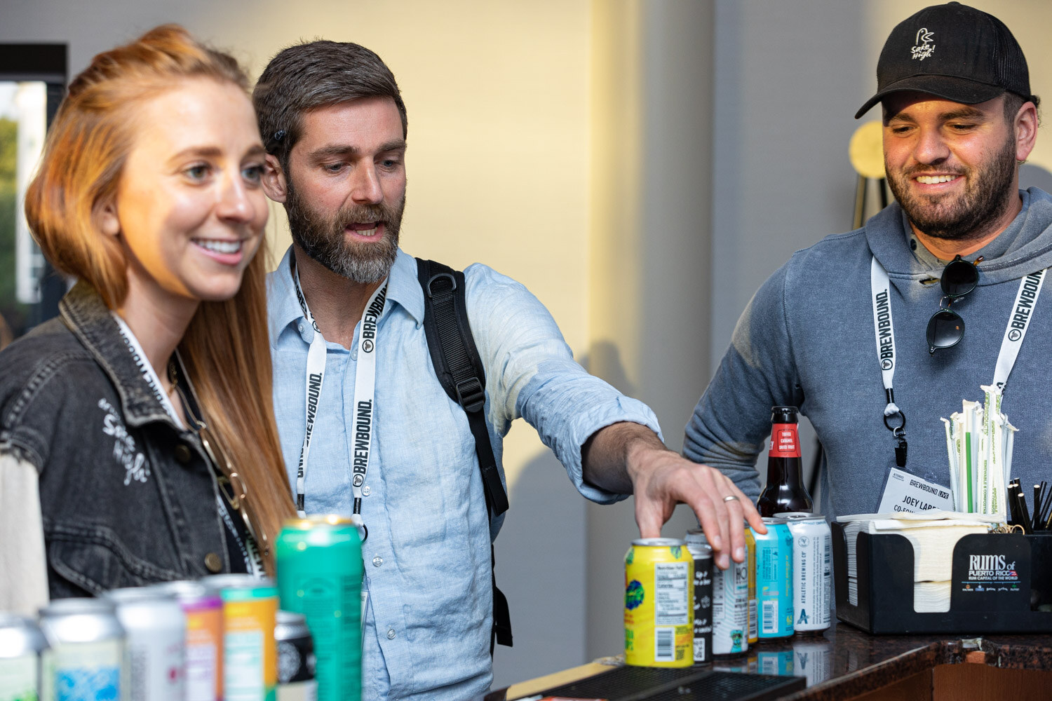 A woman and two men smile as they reach toward a bar lined with beverage cans.