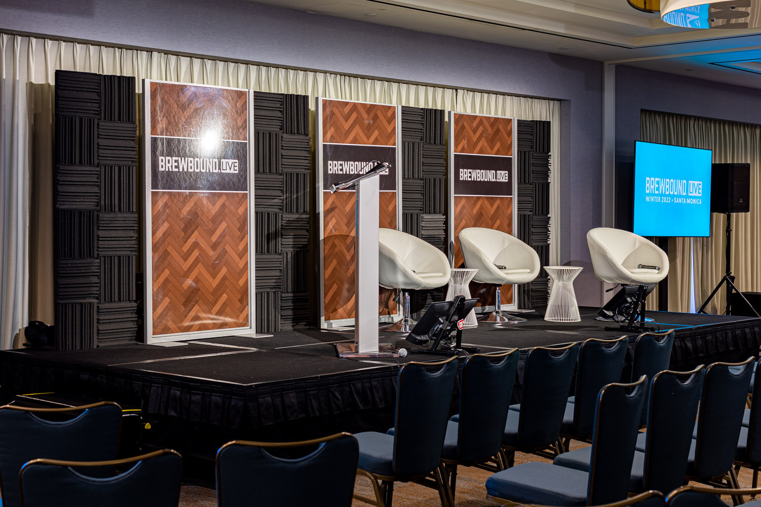 A stage with three white scoop chairs, a white podium and Brewbound Live signs on dark acoustic panels, with empty seats in the foreground.