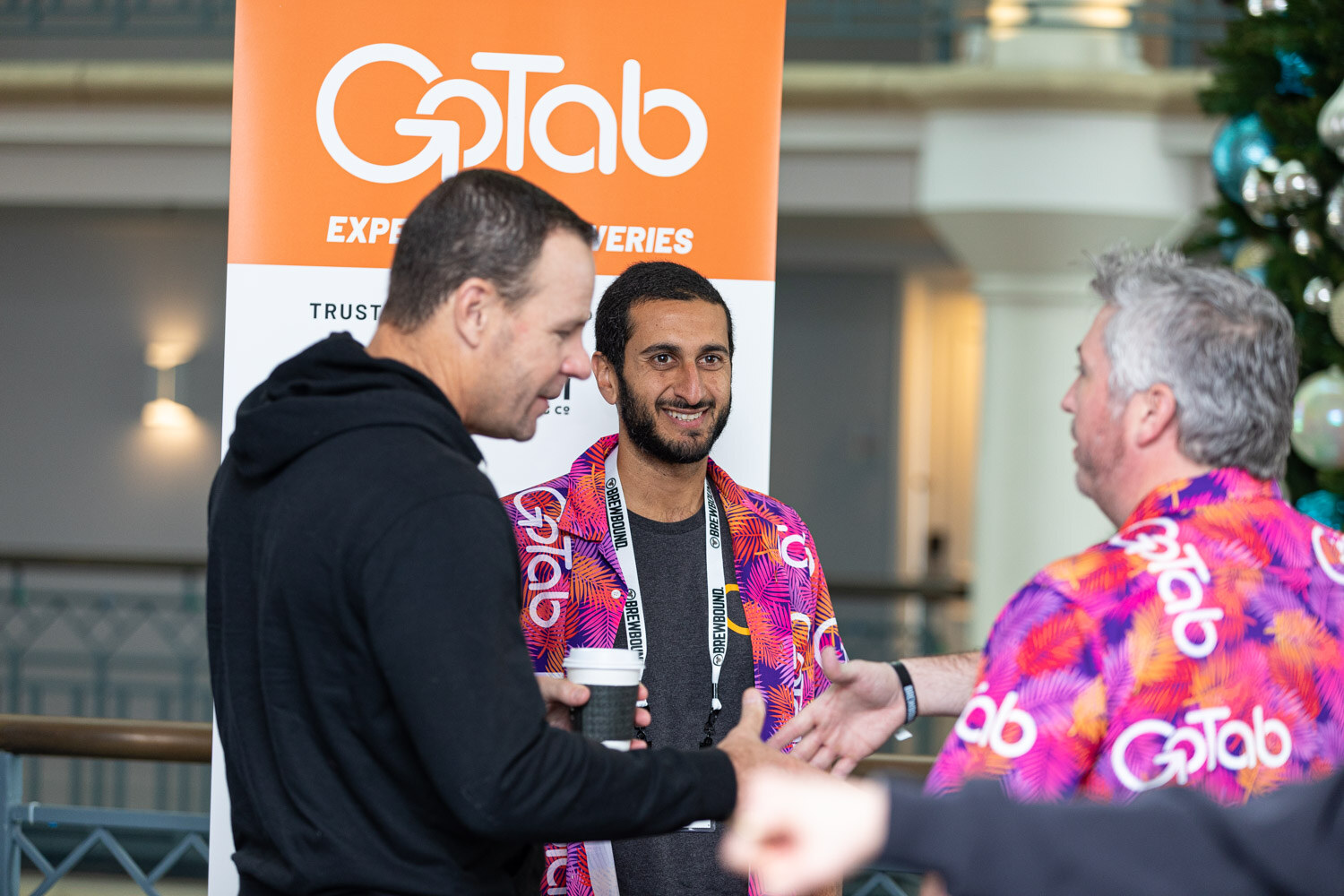 Three men talk in front of an orange GoTab banner, two of them in bright pink and purple GoTab-print shirts.