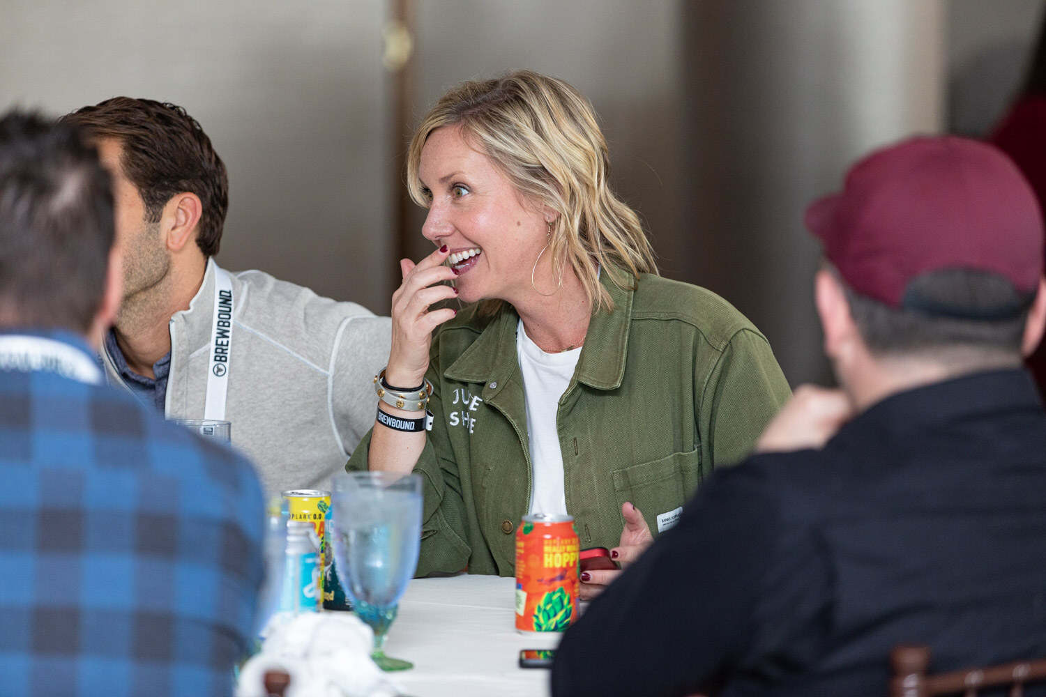 A smiling woman with blonde hair in an olive shirt-jacket sits at a round table with water glasses and cans.