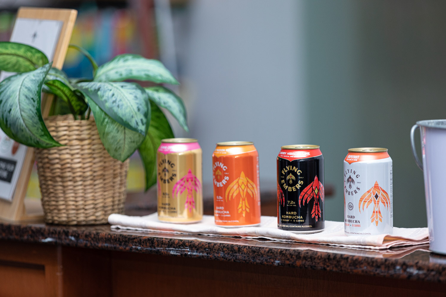 Four colorful beverage cans lined up on a wooden counter next to a basket with a leafy plant.