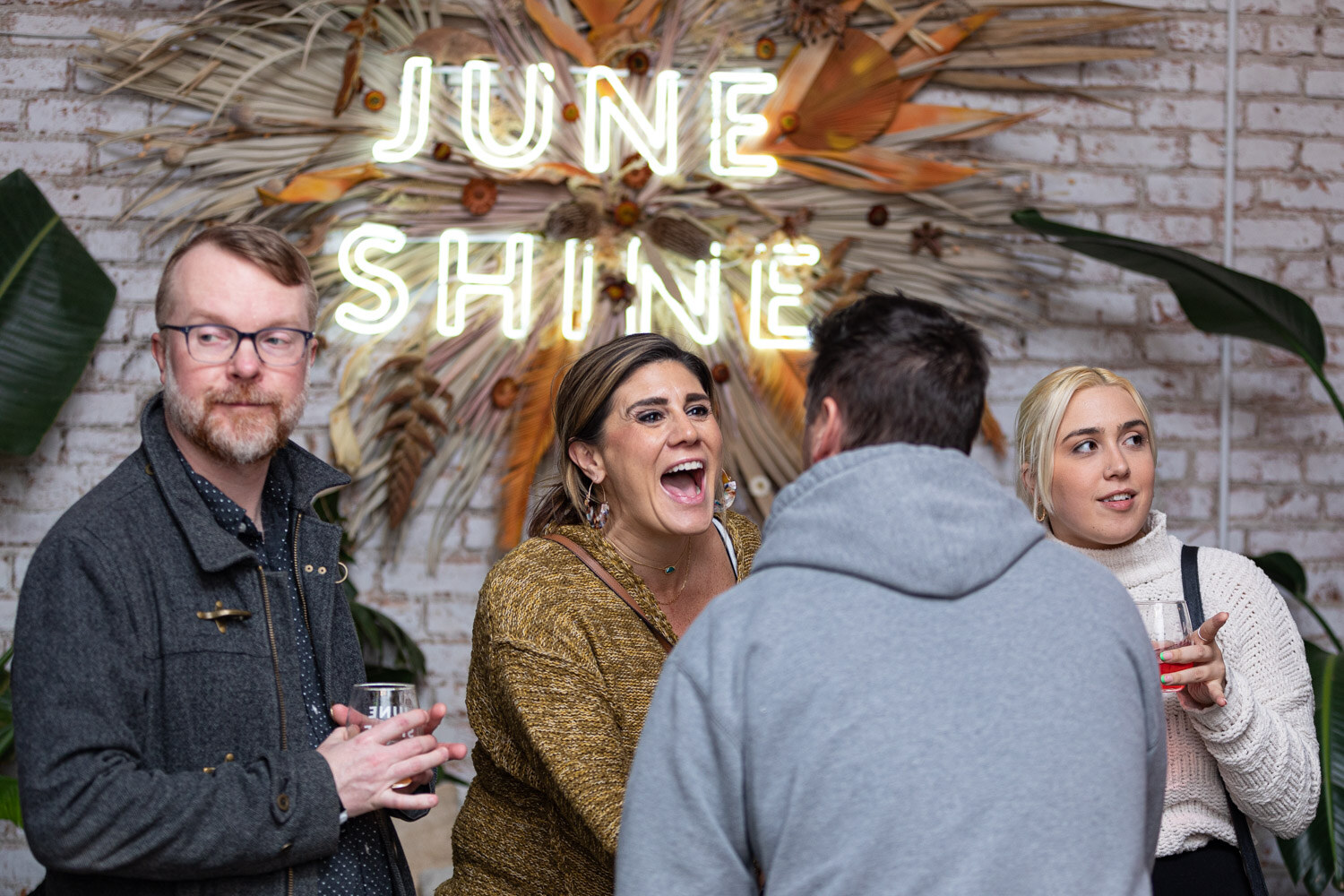 A man with glasses, a laughing woman in a mustard sweater and others stand under a lit JuneShine neon sign on a white brick wall.