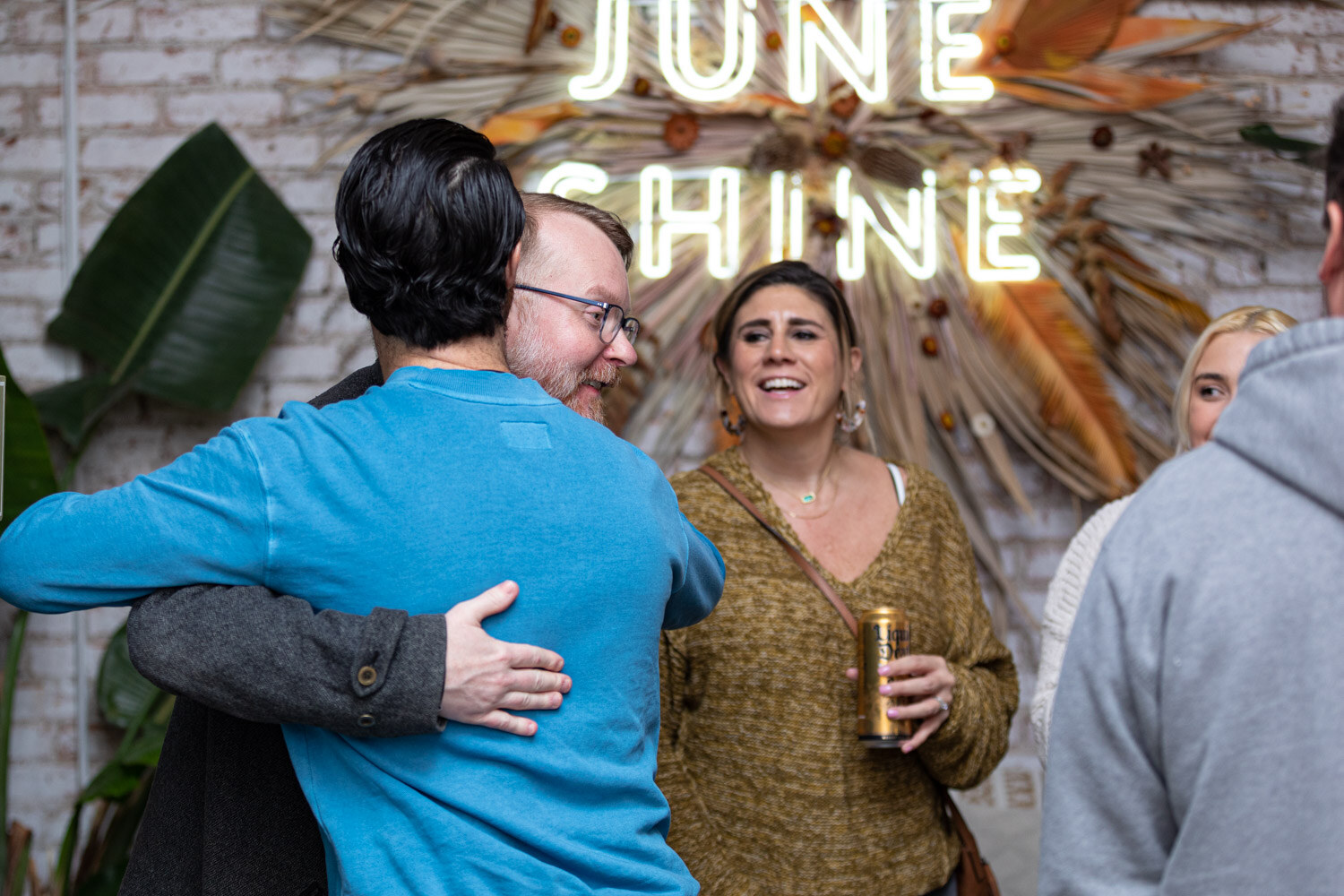 A man in a bright blue sweater hugs a bearded man in glasses beneath the June Shine neon sign while a laughing woman holds a can.