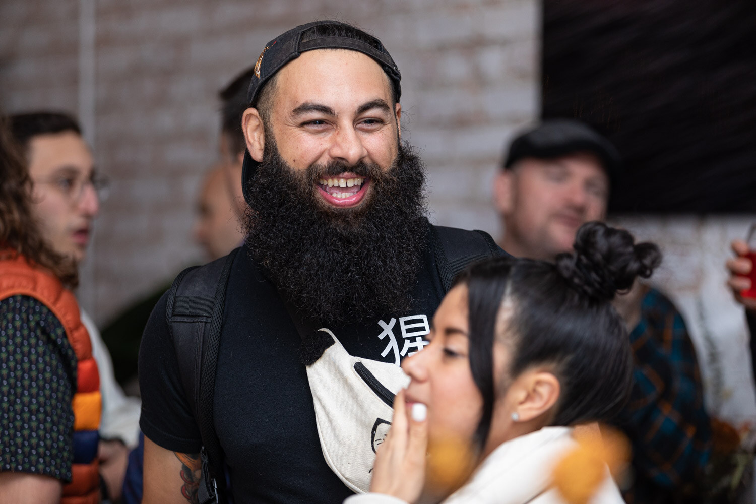 A man with a long beard and a backward black cap laughs broadly beside a woman holding her hand to her mouth.