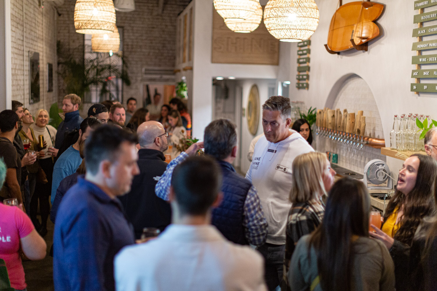 Crowded room of guests standing around a bar with beer taps, white brick walls and woven pendant lamps.
