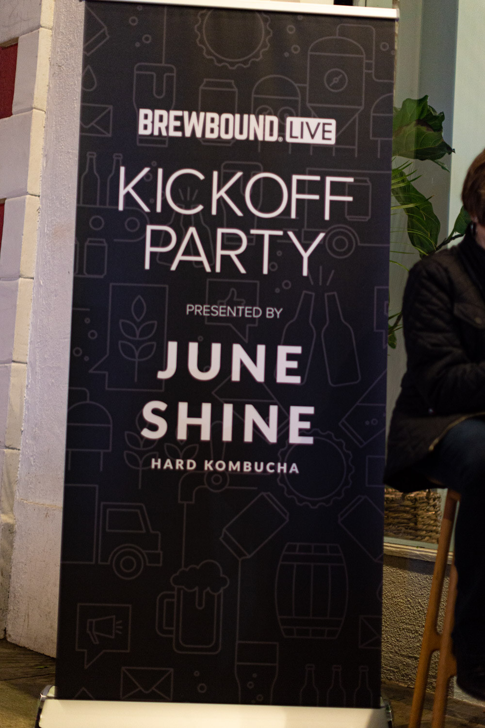A black standing banner reading Brewbound Live Kickoff Party presented by June Shine Hard Kombucha next to a brick wall.
