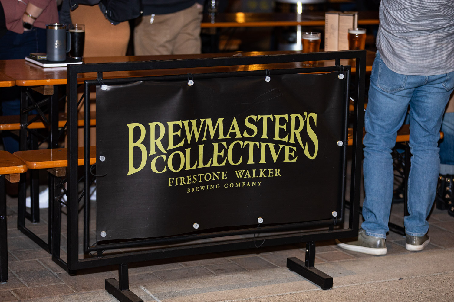 A black banner reading Brewmaster's Collective Firestone Walker Brewing Company on a metal stand with picnic tables behind it at night.
