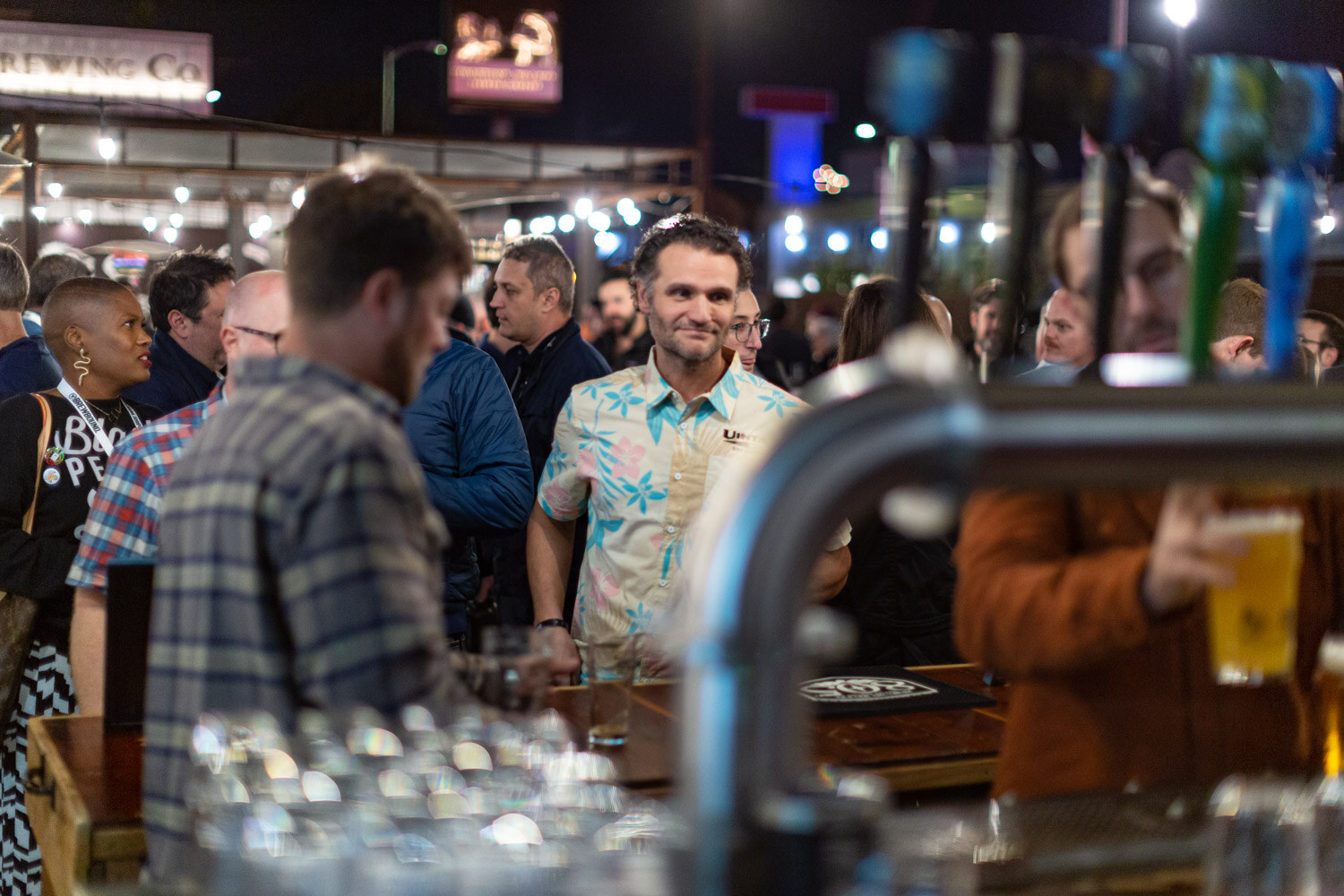 A busy bar with beer tap handles in the foreground and guests with lanyards gathered behind it under string lights at night.