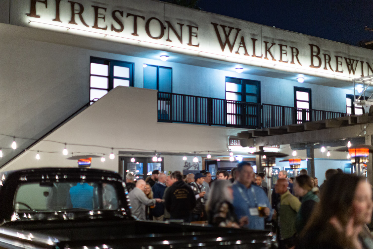 The Firestone Walker Brewing facade lit at night with a black classic truck in the foreground and a crowd behind it.
