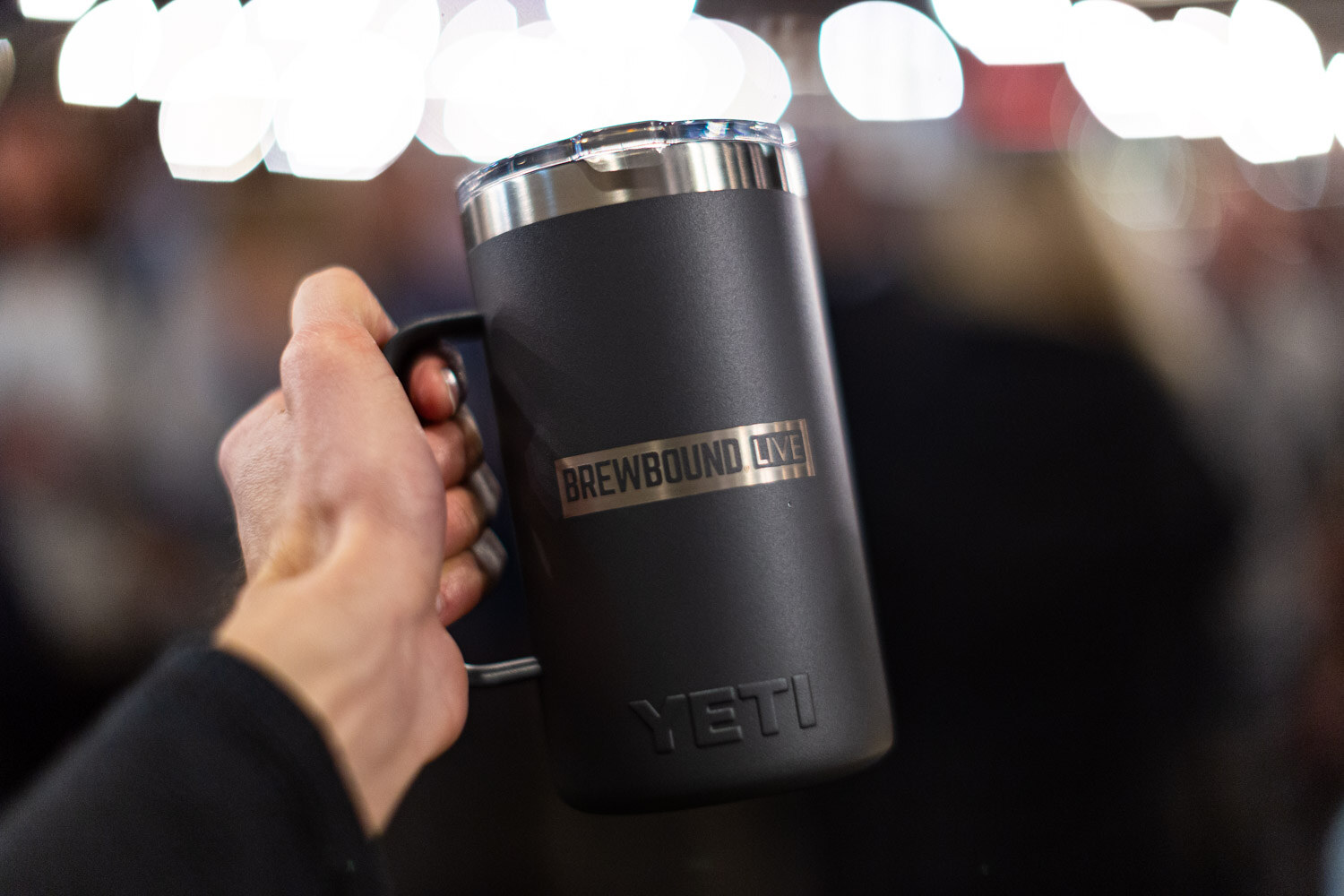 A hand raising a dark gray YETI mug engraved with Brewbound Live in front of blurred party lights.