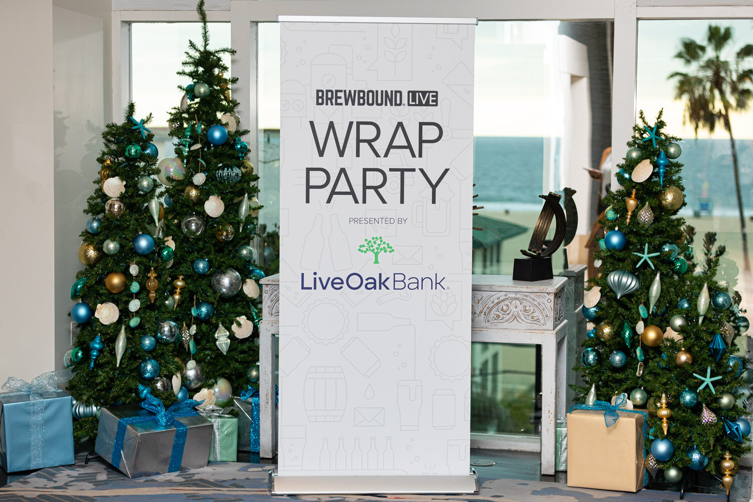 A white banner reading Brewbound Live Wrap Party presented by Live Oak Bank between two decorated Christmas trees with an ocean view behind.