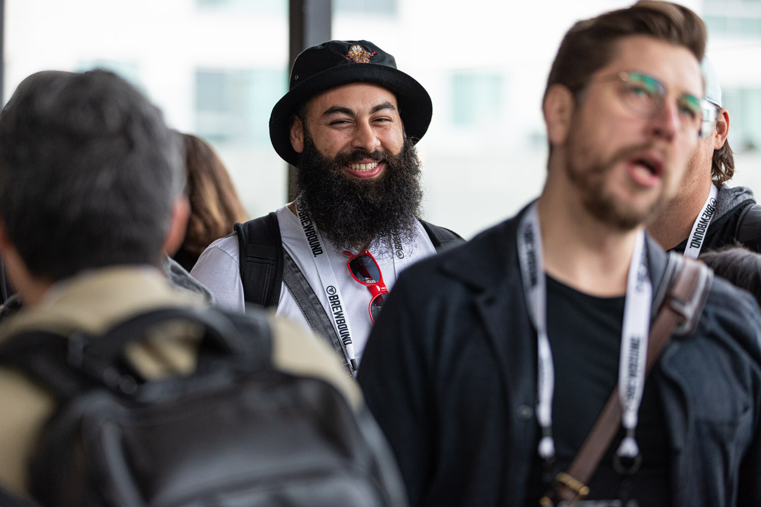 A bearded man in a black bucket hat with sunglasses on his collar laughs beside a man in a dark jacket with a lanyard.