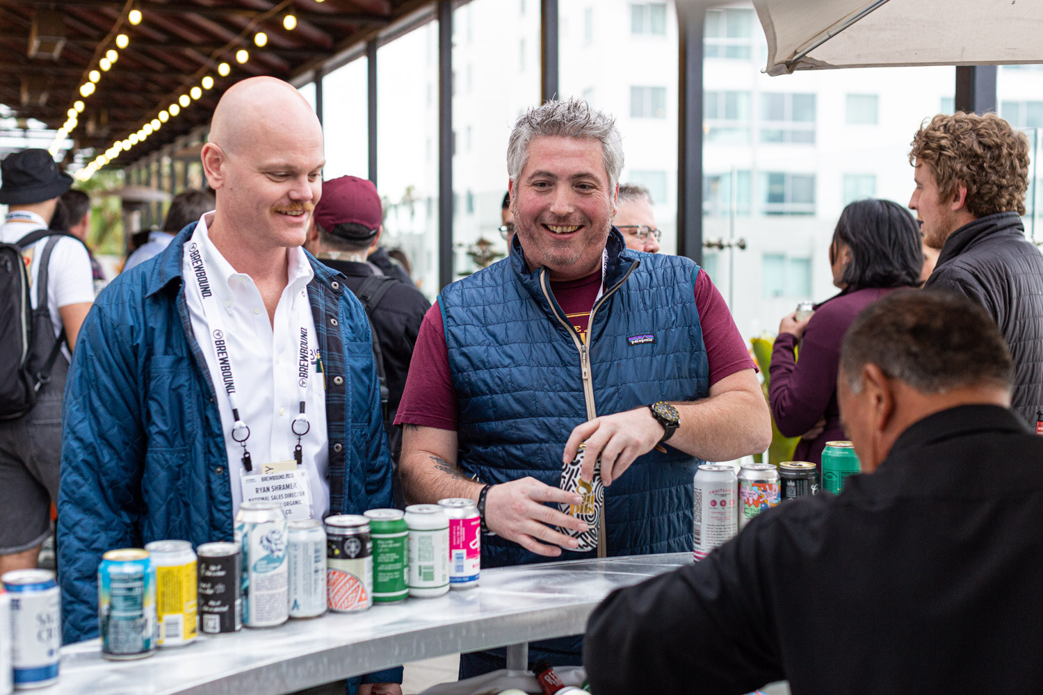 A bald man in a blue jacket and a man in a navy vest laugh beside a counter lined with colorful cans.