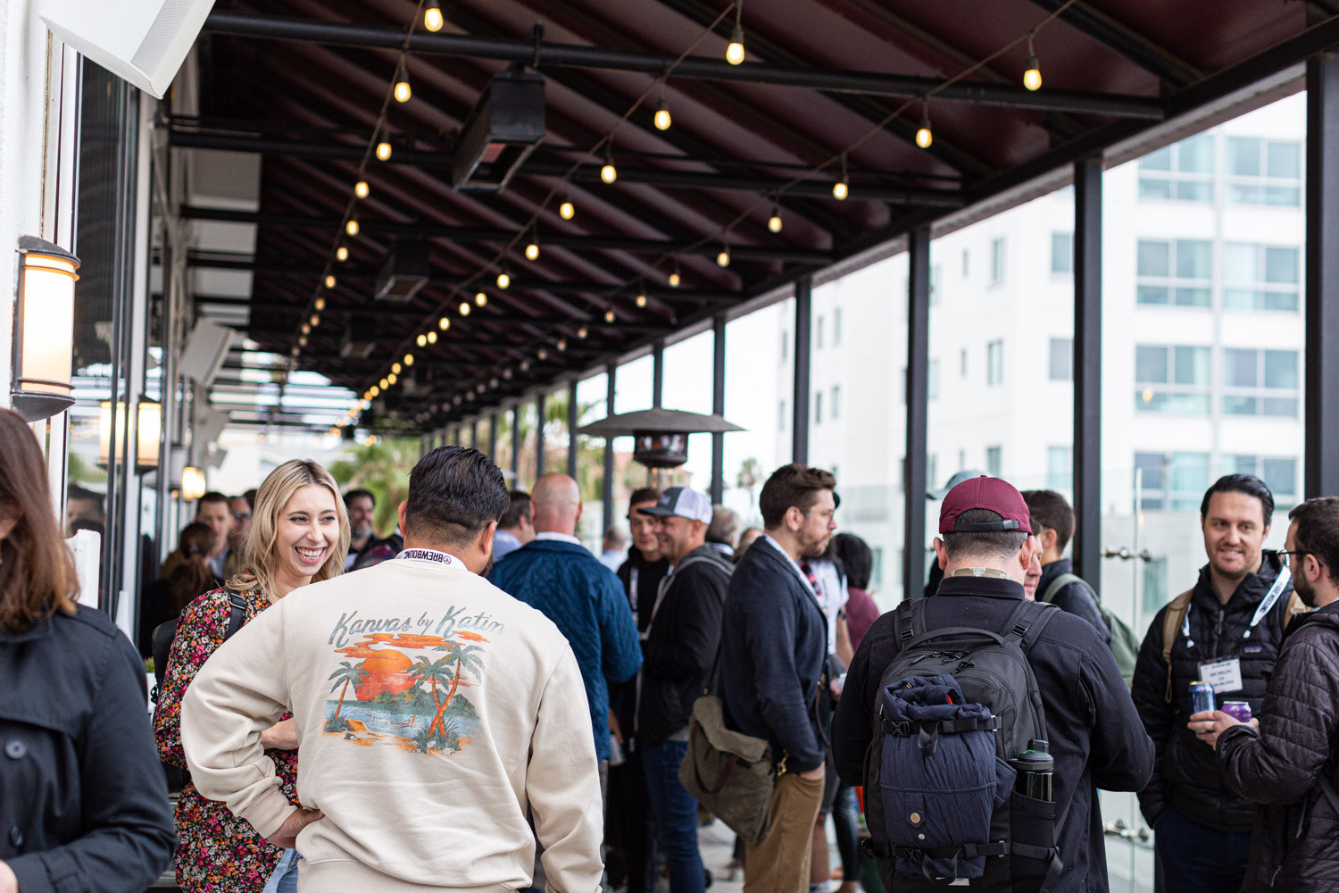 A crowd of attendees on a covered terrace with string lights and patio heaters, several in matching cream sweatshirts.